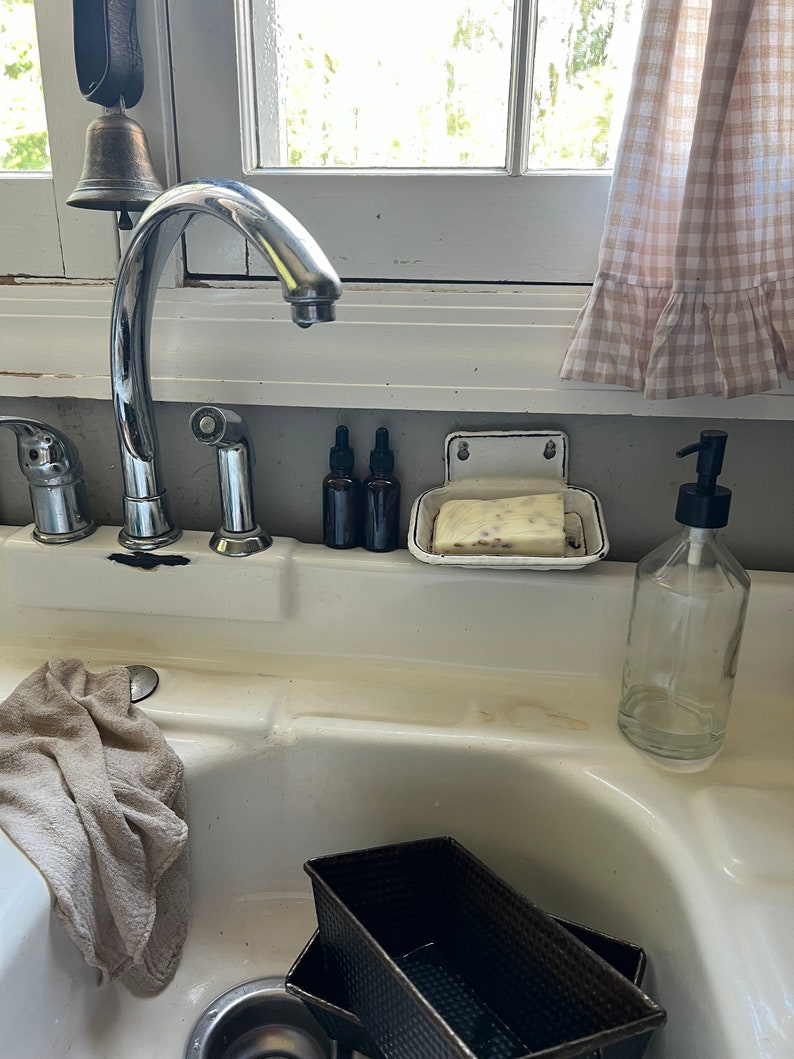 K&ouml;nnte beinhalten: Ein K&uuml;chenbecken mit einer verchromten Armatur, einer Seifenschale und zwei dunklen Glasflaschen. Ein klarer Glas-Seifenspender und zwei schwarze Brotformen befinden sich im Waschbecken. Ein beiges Handtuch h&auml;ngt an der Seite des Waschbeckens.