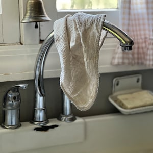 K&ouml;nnte beinhalten: Nahaufnahme eines K&uuml;chenbeckens mit einer verchromten Armatur und einem cremefarbenen Tuch, das am Auslauf h&auml;ngt. &Uuml;ber der Armatur h&auml;ngt eine Messingglocke. Ein St&uuml;ck Seife liegt in einer wei&szlig;en Seifenschale.