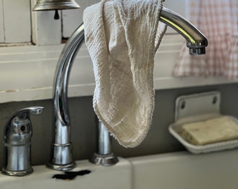 May include: A close-up of a kitchen sink with a chrome faucet and a cream-colored cloth hanging from the spout. A brass bell hangs above the faucet. A bar of soap sits in a white soap dish.