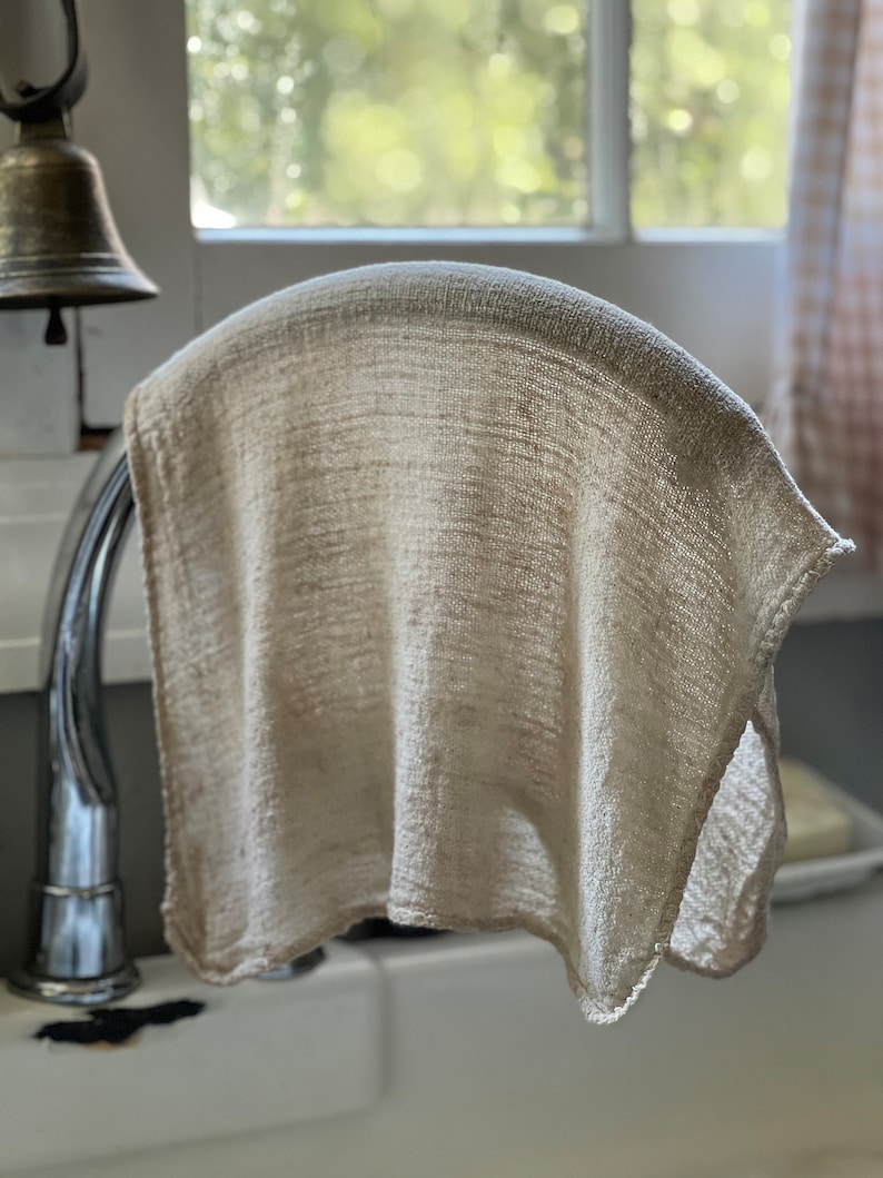 K&ouml;nnte beinhalten: Ein beiges, gewebtes K&uuml;chentuch, das &uuml;ber einem verchromten Wasserhahn drapiert ist. Das Tuch hat ein strukturiertes Aussehen und h&auml;ngt vor einem Fenster. Im Hintergrund ist eine kleine Messingglocke zu sehen.