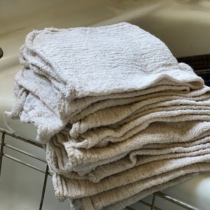 K&ouml;nnte beinhalten: Ein Stapel beiger, strukturierter Waschlappen. Die T&uuml;cher sind gefaltet und gestapelt, mit sichtbaren Rippen und einer leicht unebenen Textur. Der Stapel liegt auf einem Metallgestell in der N&auml;he eines wei&szlig;en Waschbeckens.