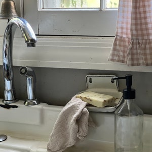 K&ouml;nnte beinhalten: Ein wei&szlig;es Porzellanwaschbecken mit einer verchromten Armatur, einer Seifenschale und einer durchsichtigen Glasflasche mit schwarzer Pumpe. Ein St&uuml;ck Seife und ein beiges Waschlappen befinden sich im Waschbecken. Im Hintergrund ein Fenster mit kariertem Vorhang.