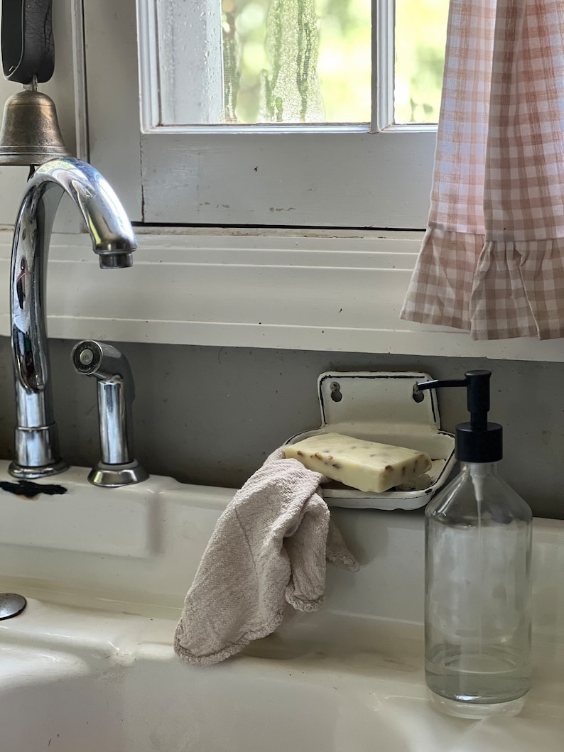 K&ouml;nnte beinhalten: Ein wei&szlig;es Porzellanwaschbecken mit einer verchromten Armatur, einer Seifenschale und einer durchsichtigen Glasflasche mit schwarzer Pumpe. Ein St&uuml;ck Seife und ein beiges Waschlappen befinden sich im Waschbecken. Im Hintergrund ein Fenster mit kariertem Vorhang.