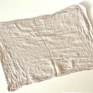 K&ouml;nnte beinhalten: Ein rechteckiges, beiges Leinen-Servietten mit rohem Rand. Der Stoff hat eine zerknitterte Textur, und die Kanten sind leicht ausgefranst. Die Serviette liegt flach auf einer wei&szlig;en Oberfl&auml;che und zeigt ihr schlichtes, rustikales Design.