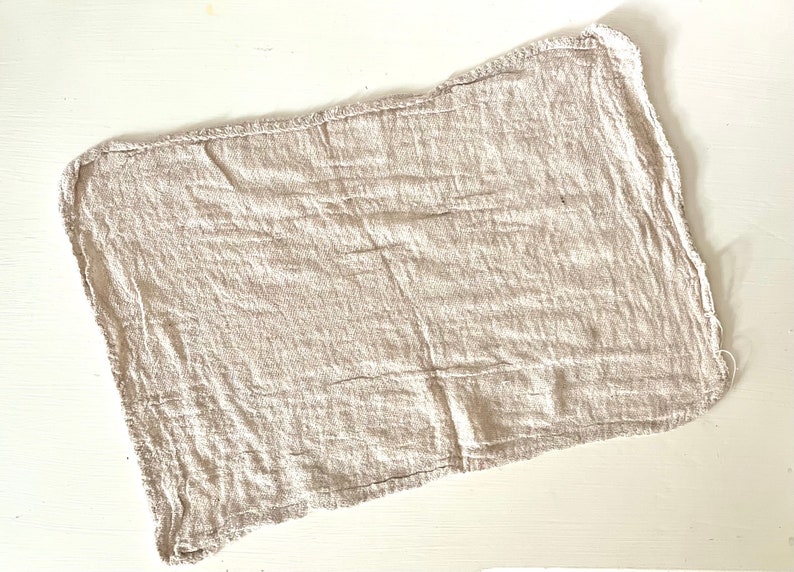 K&ouml;nnte beinhalten: Ein rechteckiges, beiges Leinen-Servietten mit rohem Rand. Der Stoff hat eine zerknitterte Textur, und die Kanten sind leicht ausgefranst. Die Serviette liegt flach auf einer wei&szlig;en Oberfl&auml;che und zeigt ihr schlichtes, rustikales Design.