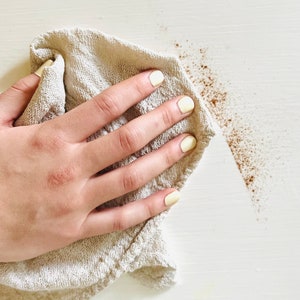 K&ouml;nnte beinhalten: Eine Hand mit gelbem Nagellack h&auml;lt ein beiges Tuch und wischt eine wei&szlig;e Oberfl&auml;che ab. Braune Partikel sind auf der Oberfl&auml;che verstreut, was auf eine Reinigung hindeutet. Das Tuch ist strukturiert, und der Hintergrund ist einfarbig wei&szlig;.