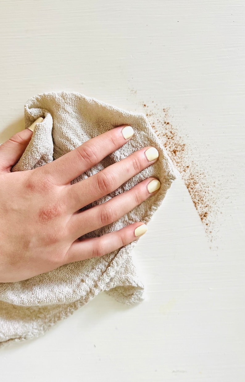K&ouml;nnte beinhalten: Eine Hand mit gelbem Nagellack h&auml;lt ein beiges Tuch und wischt eine wei&szlig;e Oberfl&auml;che ab. Braune Partikel sind auf der Oberfl&auml;che verstreut, was auf eine Reinigung hindeutet. Das Tuch ist strukturiert, und der Hintergrund ist einfarbig wei&szlig;.