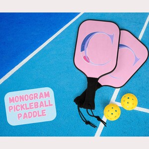 Könnte beinhalten: Zwei rosa Pickleball-Schläger mit einem Monogramm "C", schwarzen Griffen und zwei gelben Pickleballs auf einem blauen Platz. Der Text "MONOGRAM PICKLEBALL PADDLE" befindet sich in einer weißen Box.