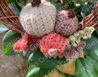 May include: A crocheted autumn wreath with a brown twig base, featuring a white pumpkin, a brown and green acorn, and a red and brown apple. The wreath is hanging on a green leafy branch.