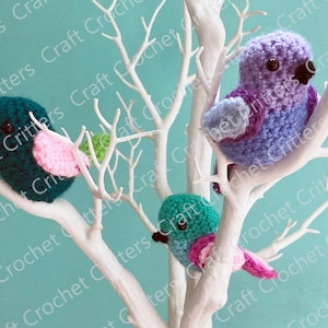May include: Three crocheted birds perched on a white branch. The birds are teal with pink accents, purple with blue accents, and teal with pink accents. The birds are on a white branch against a turquoise background.