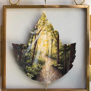 May include: A dried leaf painted with a forest scene featuring a sunlit path between trees, encased in a square brass frame with a hanging loop. The painting depicts light rays filtering through foliage, with the initials MB in gold paint at the bottom.
