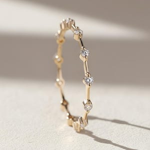 May include: A delicate gold ring featuring a series of small, sparkling diamonds. The ring's design incorporates a linear pattern with evenly spaced gemstones, creating a minimalist yet elegant aesthetic. The ring is shown on a neutral background, highlighting its fine details.