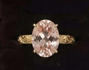 May include: A gold ring featuring a large, oval-cut, pale peach-coloured gemstone set in a four-prong mounting. The gold band displays an intricate, engraved scrollwork pattern along its surface, presented inside a dark brown wooden box.