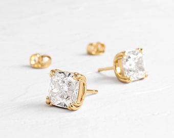 May include: Close-up of two gold stud earrings with large, square, clear gemstones. The earrings are set in a gold frame with a textured design. Two additional gold earring backs are visible.