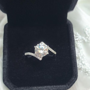 May include: A silver engagement ring with a large, round, clear gemstone. The ring is set in a twisted band with smaller accent stones. The ring is displayed in a black velvet ring box.