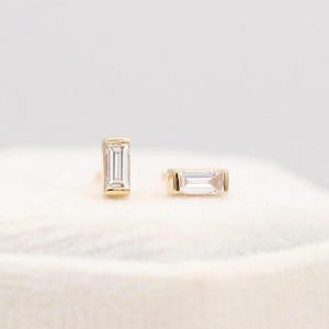 Puede incluir: Un par de pendientes de oro con gemas rectangulares y transparentes. Los pendientes están engastados en un engaste de bisel dorado y se muestran sobre una superficie blanca texturizada. Las gemas tienen un corte baguette.