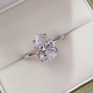 Puede incluir: Anillo de compromiso solitario con un diamante en forma de pera engastado en una fina banda plateada. El diamante claro y facetado refleja la luz. El anillo se presenta en un joyero verde claro.