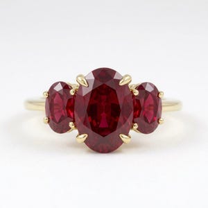 May include: A three-stone ring featuring a large oval-cut deep red gemstone in the center, flanked by two smaller oval-cut red gemstones. The stones are set in a polished yellow gold band with delicate prongs securing each gem.