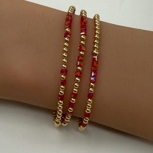 Puede incluir: Tres pulseras doradas con cuentas cuadradas rojas alternas. Las pulseras están envueltas alrededor de una muñeca, mostrando el diseño y el contraste de color de la joyería. Las cuentas doradas son pequeñas y redondas.