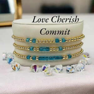 Op de afbeelding: Vier gouden armbanden met turquoise blauwe kralen. De armbanden zijn gestapeld op een witte displaystandaard. De tekst "Love Cherish Commit" staat boven de armbanden.