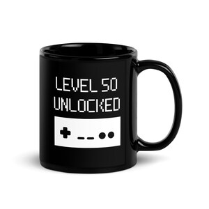 May include: Black ceramic coffee mug with white text that reads "LEVEL 50 UNLOCKED" and a pixelated image of a video game controller.