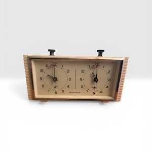 May include: A vintage chess clock with two circular clock faces. The clock has a light wood-toned frame with a rectangular shape. Each clock face displays black numerals and hands, with a red accent at the top. The clock has two black buttons on top.