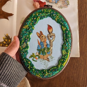 May include: Embroidered hoop art featuring Peter Rabbit in a blue jacket, holding a carrot, with a red robin perched on a branch. The background is a light blue with green foliage around the edge.