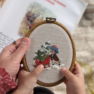 May include: A hand-stitched embroidery hoop with a white background featuring a small rabbit pulling a red sled with a Christmas tree. The rabbit is wearing a blue hat and a brown coat. The embroidery is framed in a natural wood hoop.