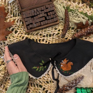 Könnte beinhalten: Schwarzer Kragen mit weißer Spitzenborte und einem handgestickten Eichhörnchen-Design. Das Eichhörnchen ist von grünem Stechpalmenlaub und roten Beeren umgeben. Ein schwarzes Samtband ist am Ausschnitt gebunden.