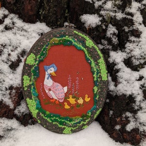 May include: Embroidered hoop art featuring a mother duck with her ducklings. The duck is wearing a blue bonnet and a pink dress. The ducklings are yellow and are surrounded by green and pink flowers. The hoop is surrounded by a green and blue border.