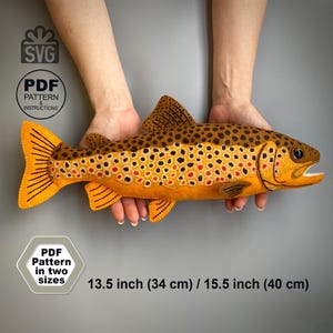 May include: A felt brown trout fish toy featuring dark brown spots and orange accents. Text includes SVG, PDF PATTERN & INSTRUCTIONS, and PDF Pattern in two sizes 13.5 inch (34 cm) / 15.5 inch (40 cm). The fish has a detailed tail and fins.