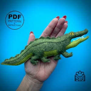 May include: Green felt alligator plush toy with a yellow eye and open mouth. The alligator is being held in a hand. The image also includes the text "PDF PATTERN & INSTRUCTIONS" and "SVG".