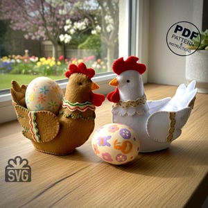 Peut inclure: Deux paniers de Pâques en forme de poule en feutre, un marron et un blanc, chacun contenant un œuf décoré. La poule marron a une crête et des barbillons rouges, et la poule blanche a un col en dentelle. Un œuf décoré supplémentaire est devant.