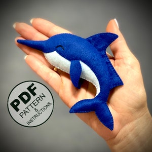 May include: A handmade blue felt dolphin with white accents, featuring a long snout and a curved tail. The dolphin is approximately 6 inches long. The image includes a circular graphic with the text "PDF PATTERN & INSTRUCTIONS".