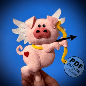 Puede incluir: Un cerdo rosa de fieltro con alas, sosteniendo una flecha amarilla y negra. El cerdo tiene un corazón rojo en un lado y un halo amarillo. El texto "PDF PATTERN & INSTRUCTIONS" está en un círculo blanco con un borde negro.