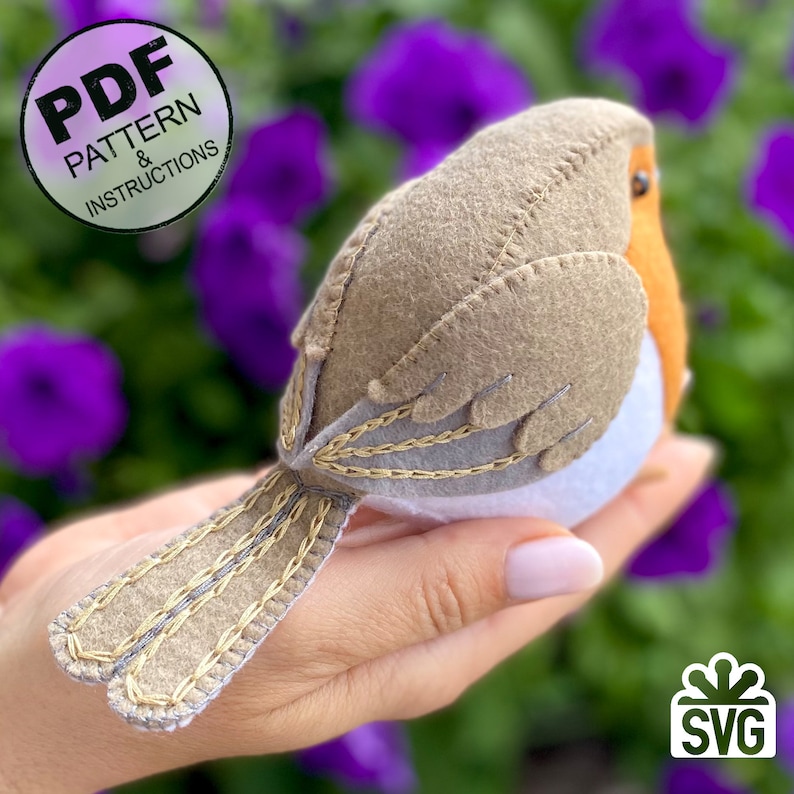 K&ouml;nnte beinhalten: Ein Filz-Rotkehlchen mit braunem K&ouml;rper, wei&szlig;er Brust und orangefarbener Brust. Der Vogel hat einen langen Schwanz mit goldenen und grauen N&auml;hten. Der Vogel wird in einer Hand vor einem Hintergrund aus lila Blumen gehalten.