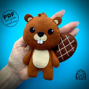 May include: A handmade brown felt beaver plushie with a beige belly and a brown tail. The beaver has black eyes, a black nose, and two white teeth. The text "PDF PATTERN & INSTRUCTIONS" is in a circle on the left side of the image.