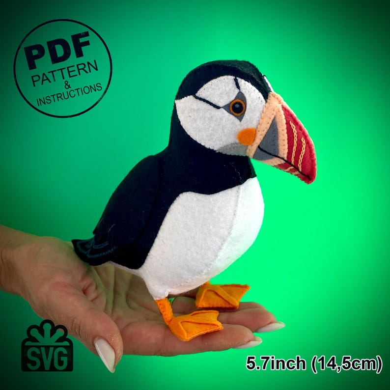 Peut inclure: Un jouet en peluche de macareux en feutre avec des plumes noires et blanches et un bec orange. Le jouet mesure 14,5 cm de haut et est tenu dans la main d'une personne. L'image comprend le texte "PDF PATTERN & INSTRUCTIONS" et "SVG".