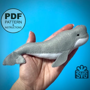 May include: A gray felt whale plush toy with white felt detailing. The whale is being held in a hand against a blue background. The image also includes text that says "PDF PATTERN & INSTRUCTIONS" and "SVG".