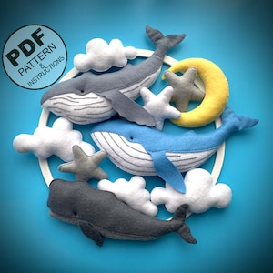 May include: A felt whale mobile with a white hoop, clouds, stars, and a crescent moon. The whales are grey and blue with white stitching. The text "PDF PATTERN & INSTRUCTIONS" is on a circle.