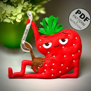 Pode incluir: Um brinquedo de pelúcia de morango vermelho de feltro com um caule e folhas de feltro verde. O morango tem um rosto sorridente com olhos pretos e uma colher de feltro marrom na mão. O brinquedo está sentado em uma superfície cinza.  PDF PATTERN & INSTRUCTIONS