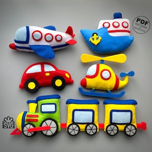 May include: A selection of colourful felt toy vehicles, including an aeroplane, boat, car, helicopter, and train. The aeroplane is white with blue and red accents. The boat is blue with a yellow anchor. The car is red with black wheels. The helicopter is yellow and red. The train is green, yellow, and blue.