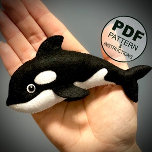 May include: A handmade felt orca whale toy in black and white, with a small eye detail. The toy is held in a hand. A circular graphic in the upper right corner reads "PDF PATTERN & INSTRUCTIONS".
