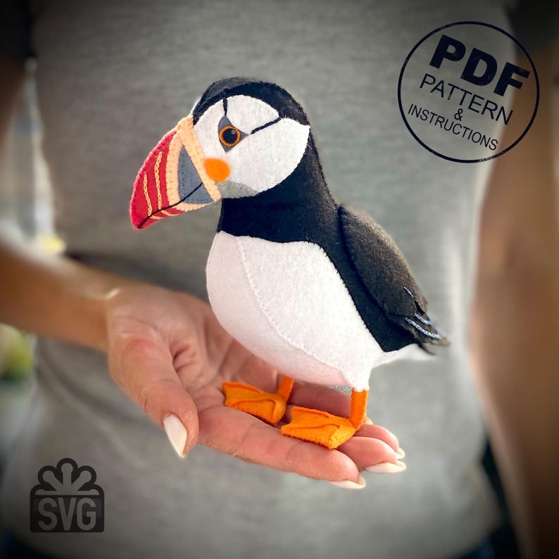 Peut inclure: Un jouet en peluche de macareux en feutre avec un corps noir et blanc, des pieds orange et un bec rouge et blanc. Le jouet est tenu dans la main d'une personne. L'image comprend &eacute;galement le texte "PDF PATTERN & INSTRUCTIONS" et "SVG".