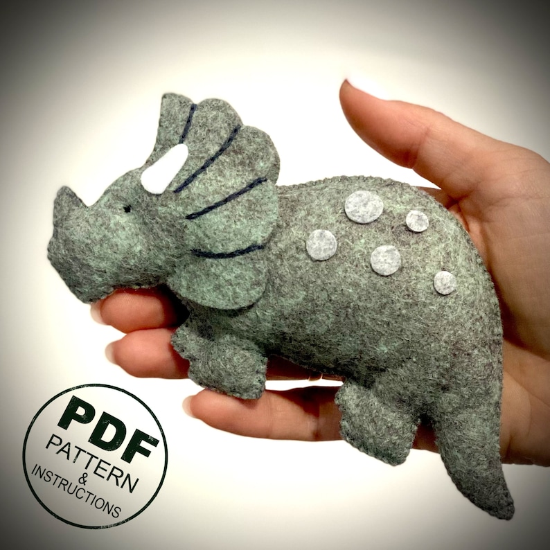 K&ouml;nnte beinhalten: Ein graues Filz-Triceratops-Pl&uuml;schtier mit wei&szlig;en Filzpunkten. Das Triceratops hat einen schwarzen Filz-Fr&auml;nschen und ein wei&szlig;es Filz-Horn. Das Pl&uuml;schtier wird in einer Hand gehalten. Das Bild enth&auml;lt auch einen gr&uuml;nen Kreis mit dem Text "PDF PATTERN & INSTRUCTIONS".