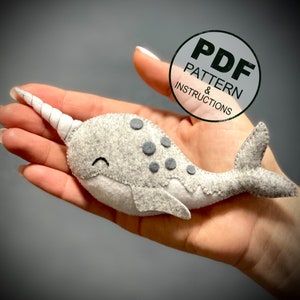May include: A gray felt narwhal plush toy with a white horn and black eyes. The toy has a smiling expression and is decorated with gray spots. The image includes the text "PDF PATTERN & INSTRUCTIONS".