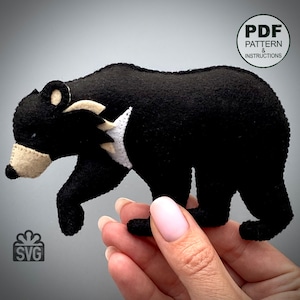 Puede incluir: Un oso de peluche negro de fieltro con detalles blancos y beige. El oso está de pie sobre una mano. La imagen incluye el texto "PDF PATTERN & INSTRUCTIONS" y "SVG".