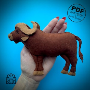 Op de afbeelding: Een bruin, gevuld knuffeldier van een buffel gemaakt van vilt met bruine hoorns en hoeven. De buffel wordt in de hand van een persoon gehouden. De afbeelding bevat ook de tekst "PDF PATTERN & INSTRUCTIONS" en "SVG".
