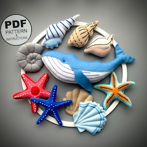 May include: A felt whale, starfish, seashells, and an ammonite are arranged on a white hoop. The felt pieces are in shades of blue, red, orange, brown, and grey. The text "PDF PATTERN & INSTRUCTIONS" is on a green label.