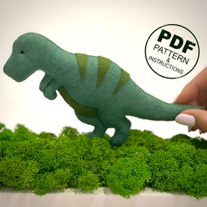 May include: A green felt dinosaur toy with a striped pattern on its back. The dinosaur is standing on a bed of green moss. The image includes the text "PDF PATTERN & INSTRUCTIONS".